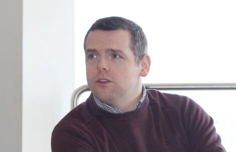 A man with short dark hair in a maroon sweater looks slightly off-camera with a neutral expression. He stands in front of a bright, windowed area with a metal railing behind him.