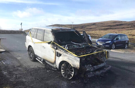 A severely burnt SUV with caution tape around it sits on a roadside with minimal traffic. Another vehicle is parked nearby, and the landscape is mostly open with hills and a cloudy sky.