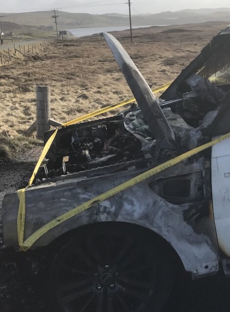 A burned-out car with an open hood is roped off with yellow caution tape. The background features rural scenery with a body of water and hills in the distance.