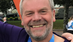A man with gray hair and a beard smiles at the camera, wearing a purple shirt and a blue race medal around his neck. Trees and an orange flag are visible in the background.