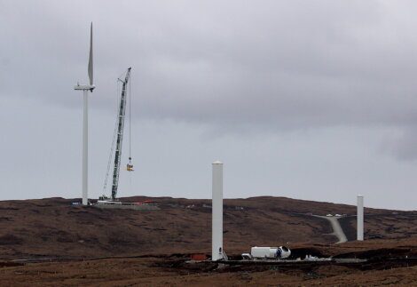 Construction of wind turbines underway in a barren, hilly landscape. One turbine is fully erected, while others are in various stages of assembly with a crane in action.