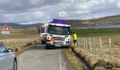 A truck with a purple roof is tilted off the road, with a person in a high-visibility jacket standing next to it. Another vehicle is on the same road, with a "No footway for 2 miles" sign visible.
