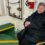 An elderly woman wearing a black coat sits on a chair in what appears to be a ship's interior. She is holding a folded black umbrella and smiling at the camera.