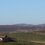 A rural landscape with a small house in the foreground and several wind turbines on rolling hills in the background.