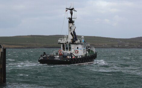 A small tugboat named "Einar" navigates through choppy waters near a coastal area with rolling hills in the background.