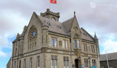 A historic stone building with a clock tower, intricate windows, and a red flag on top. The structure is identified as Lerwick Town Hall.