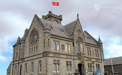 A historic stone building with a clock tower, intricate windows, and a red flag on top. The structure is identified as Lerwick Town Hall.
