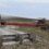 A truck transports a large wind turbine blade on a rural road, navigating a sharp turn with the blade extending over a fenced field.