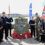 A group of people in formal attire and military uniforms stand in front of a stone memorial with wreaths and flags in the background. The setting is an outdoor location near water.