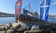 A sculpture of a boat sits on a bed of rocks with Norwegian and Scottish flags in the background under a clear blue sky.