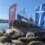 A sculpture of a boat sits on a bed of rocks with Norwegian and Scottish flags in the background under a clear blue sky.