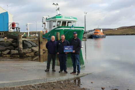 Three men standing on a pier holding a sign. A green boat and an orange boat are docked on the water in the background. The sky is cloudy.
