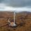 An aerial view of a single wind turbine situated in a vast, barren landscape with cloudy skies overhead. Construction equipment is visible at the base of the turbine.