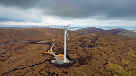 An aerial view of a single wind turbine situated in a vast, barren landscape with cloudy skies overhead. Construction equipment is visible at the base of the turbine.