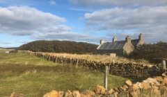 Stone cottage with a slate roof and stone walls stands in a grassy field under a partly cloudy sky.