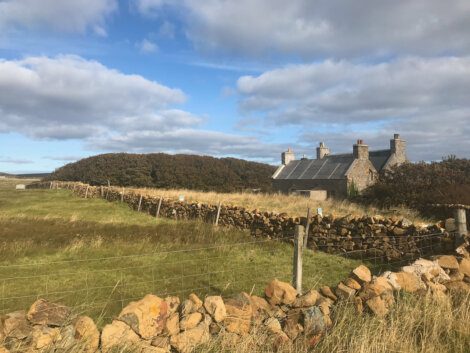 Stone cottage with a slate roof and stone walls stands in a grassy field under a partly cloudy sky.