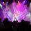 A band performs on a stage with colorful lights and fog effects. The singer at the center wears a yellow jacket, and the guitarist on the right wears a white shirt and long curly hair wig.