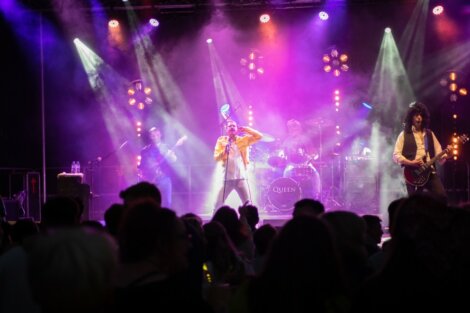 A band performs on a stage with colorful lights and fog effects. The singer at the center wears a yellow jacket, and the guitarist on the right wears a white shirt and long curly hair wig.