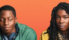 Two individuals sit against an orange background. The person on the left wears a green jacket and headphones around their neck. The person on the right wears a yellow jacket. Both look straight ahead.