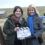Two women stand outdoors on a film set. One woman holds a clapperboard labeled "Shetland 8." Equipment and a rural landscape are visible in the background.