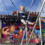 A child, strapped in a harness, is jumping on a bungee ride at a carnival with a vibrant amusement attraction in the background.