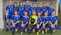 A youth girls' soccer team poses in blue uniforms for a group photo on the field in front of a building labeled "Away Team.