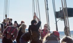 A group of people on a boat work together to raise a sail, with some observing and others taking photos. They are all wearing jackets and life vests. The sky is clear and the sea is visible in the background.
