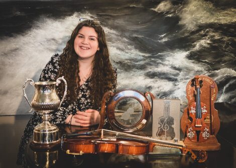 A woman stands smiling behind a table with a violin, bow, trophies, and awards, set against a backdrop of a stormy sea painting.