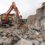 A construction excavator demolishes a partially destroyed building, with debris scattered across the site.