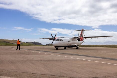 An airport worker in a high-visibility vest directs a small propeller airplane on a tarmac at an airport. The sky is partly cloudy, and there is a grassy landscape in the background.