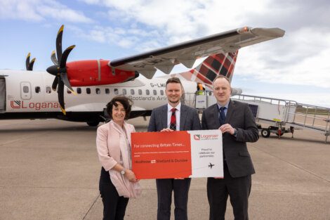 Three people stand in front of a LoganAir aircraft holding a red sign that reads, "For connecting Britain Times...." The aircraft is parked on a tarmac under a partly cloudy sky.