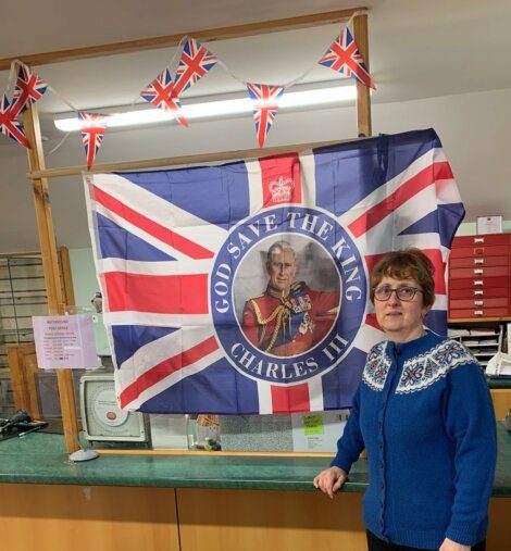 A woman stands next to a "God Save the King Charles III" flag, with Union Jack bunting above it, in a room with a green counter and various office supplies.