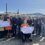 A group of people stand outdoors holding two large checks. One check is for £4,060 to COPE, and the other is for £5,100 to MS Society Scotland. Some members hold musical instruments.