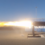 A rocket engine test is underway, emitting a bright plume of flame and exhaust on a clear day, with a shipping container in the background.
