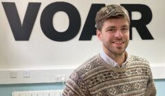 A man with short brown hair and a beard smiles while wearing a patterned sweater. He stands in front of a wall with large black letters spelling "VOAR.