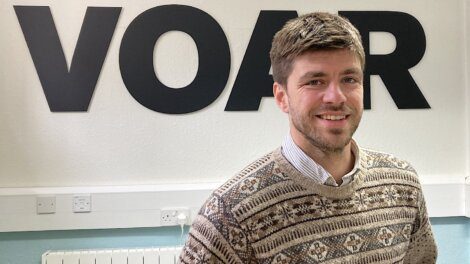 A man with short brown hair and a beard smiles while wearing a patterned sweater. He stands in front of a wall with large black letters spelling "VOAR.