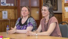 Two women sit at a table inside a room, engaging in conversation. One woman has a printed blouse and the other wears a pink top. A water bottle and various items are on the table.