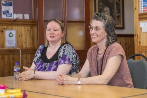 Two women sit at a table inside a room, engaging in conversation. One woman has a printed blouse and the other wears a pink top. A water bottle and various items are on the table.