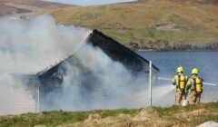 Two firefighters work to extinguish a fire in a small building near a lake, with smoke billowing out from the structure, set against a backdrop of hills and water.