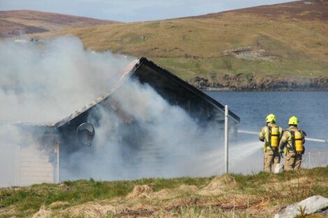 Two firefighters work to extinguish a fire in a small building near a lake, with smoke billowing out from the structure, set against a backdrop of hills and water.