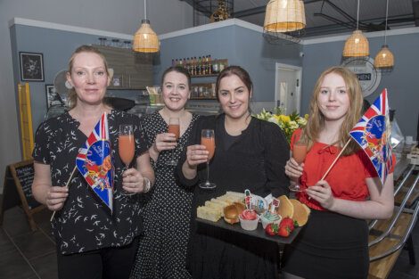 Four women stand in a restaurant, holding drinks and British flags, with a tray of snacks in front of them. The decor includes hanging lights and shelves with assorted items.