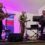 Three men play guitars and a banjo on stage in front of microphones and speakers, with a purple backdrop and a banner listing insurance and financial services.