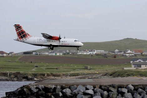 A Loganair aircraft is seen landing on a runway near the coast with houses and grassy hills in the background.