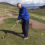 A person in a blue jacket and dark pants is preparing to swing a golf club on a golf course with rolling hills and a body of water in the background.