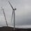 A large crane works beside a wind turbine under a cloudy sky on a barren landscape.