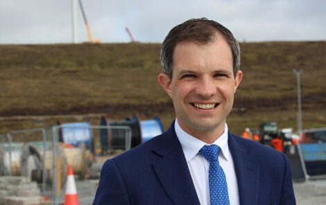 A man in a blue suit and tie smiles at the camera, standing outdoors with construction equipment and a hilly landscape in the background.