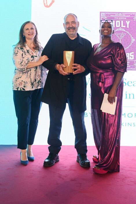 Three people standing together on a stage; the person in the middle is holding an award.