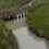 A small muddy stream flows under a short wooden bridge. Grass and dry reeds line the banks, with a wire fence running along the top.