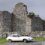 A vintage white car is parked on a road in front of an old stone castle with two round towers and a partially cloudy sky in the background.