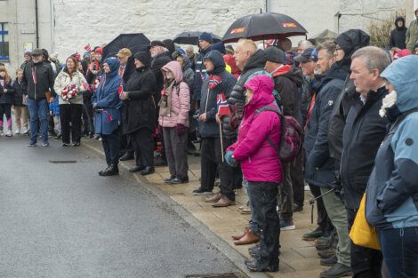 A group of people, some holding umbrellas and wearing rain gear, stand on a sidewalk in wet weather, observing an outdoor event. Some hold small flags and flowers.